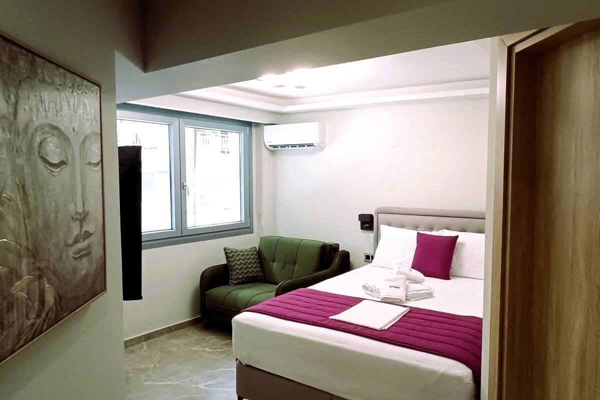 Une chambre d’hôtel avec un lit, un fauteuil vert, une fenêtre et un appareil de climatisation.