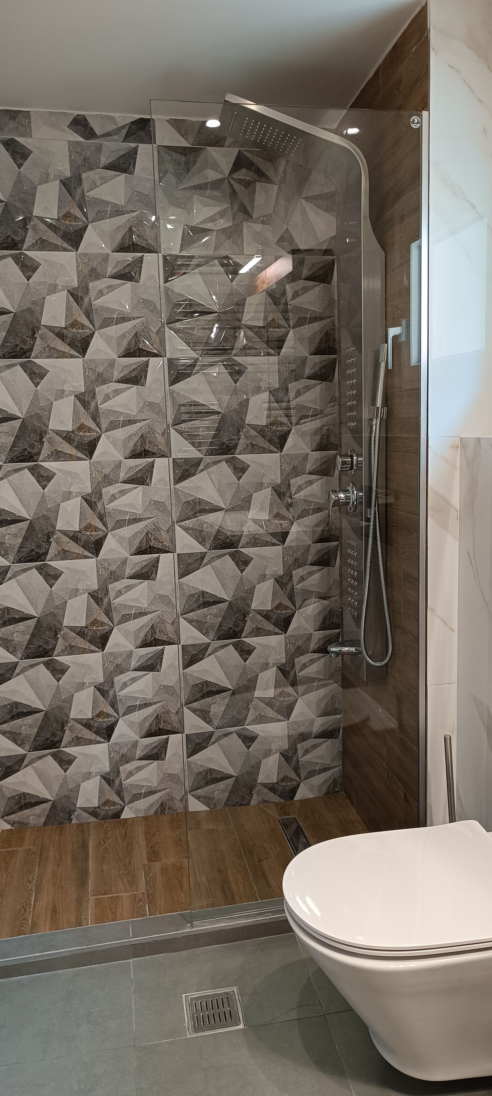 A modern bathroom with a shower enclosure featuring geometric patterned tiles, a toilet, and a wooden floor.