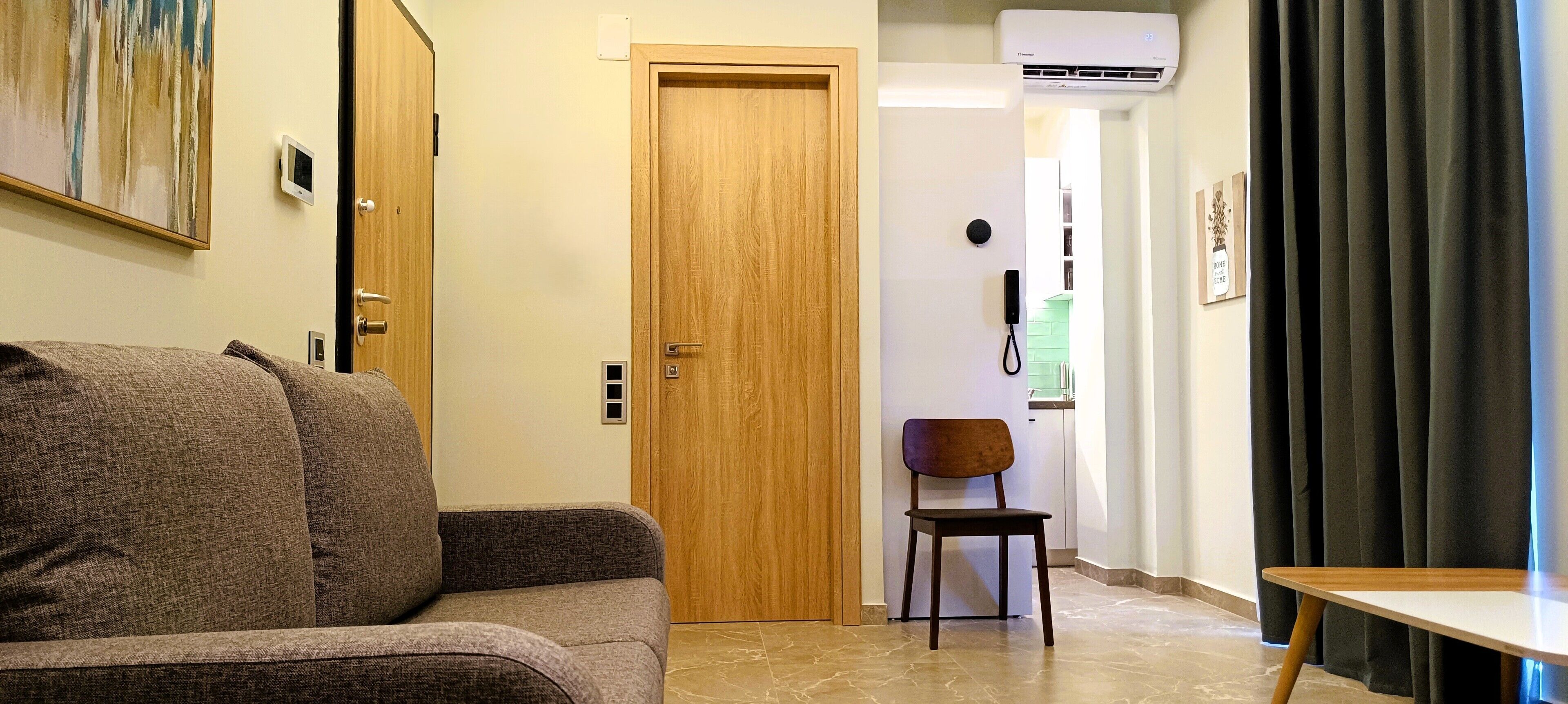 Habitación de hotel con puerta de madera, una mesita, una silla, un sofá, un cuadro en la pared y un teléfono en la pared.