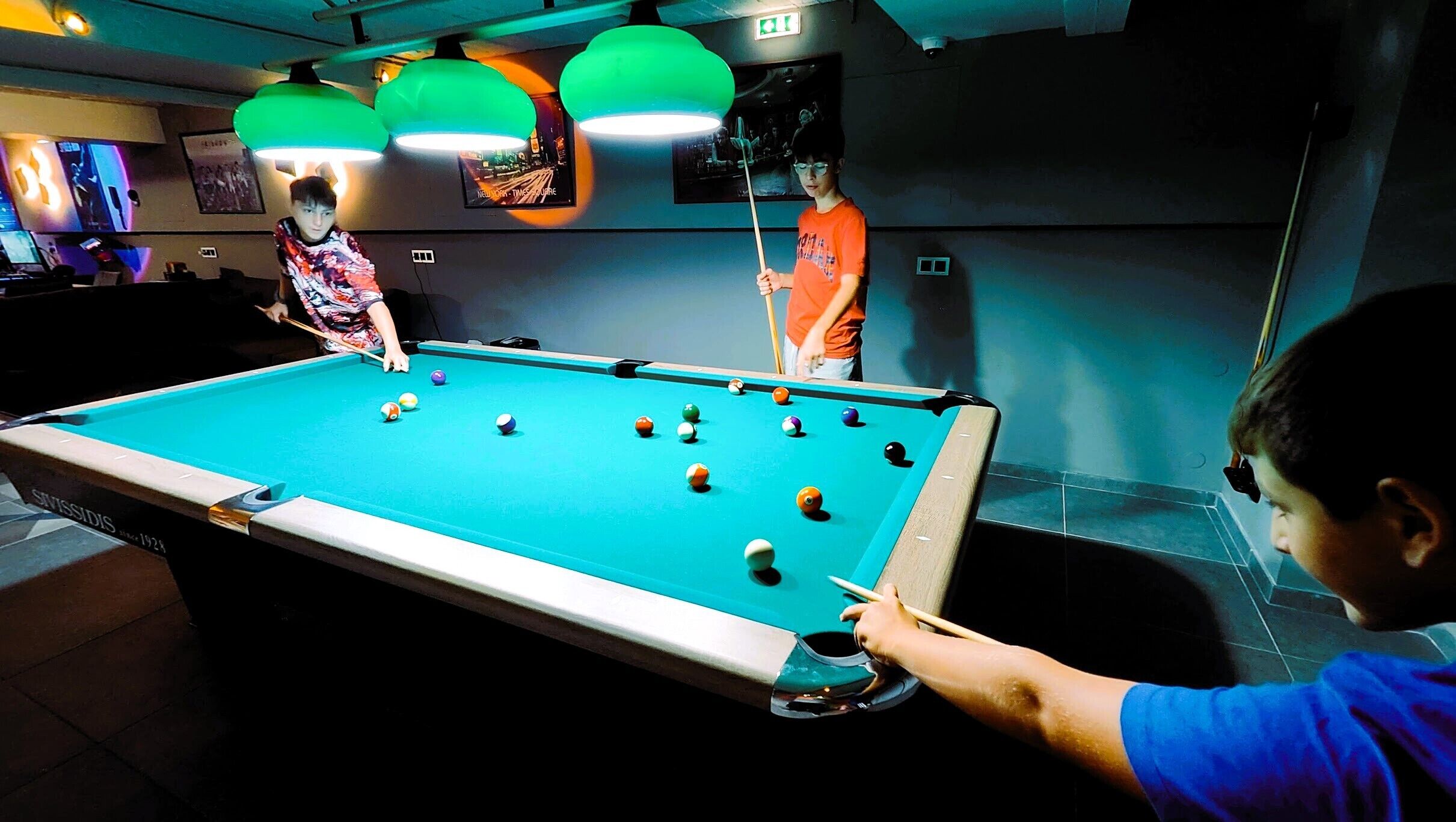 Una mesa de billar con bolas en juego, rodeada de luces verdes en el techo y una pared con un cartel.