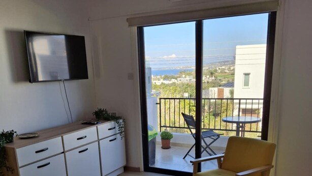 Un salon moderne avec un téléviseur à écran plat, un meuble en bois, une petite table ronde, deux chaises jaunes et une porte-fenêtre coulissante donnant sur un balcon avec vue sur la mer.
