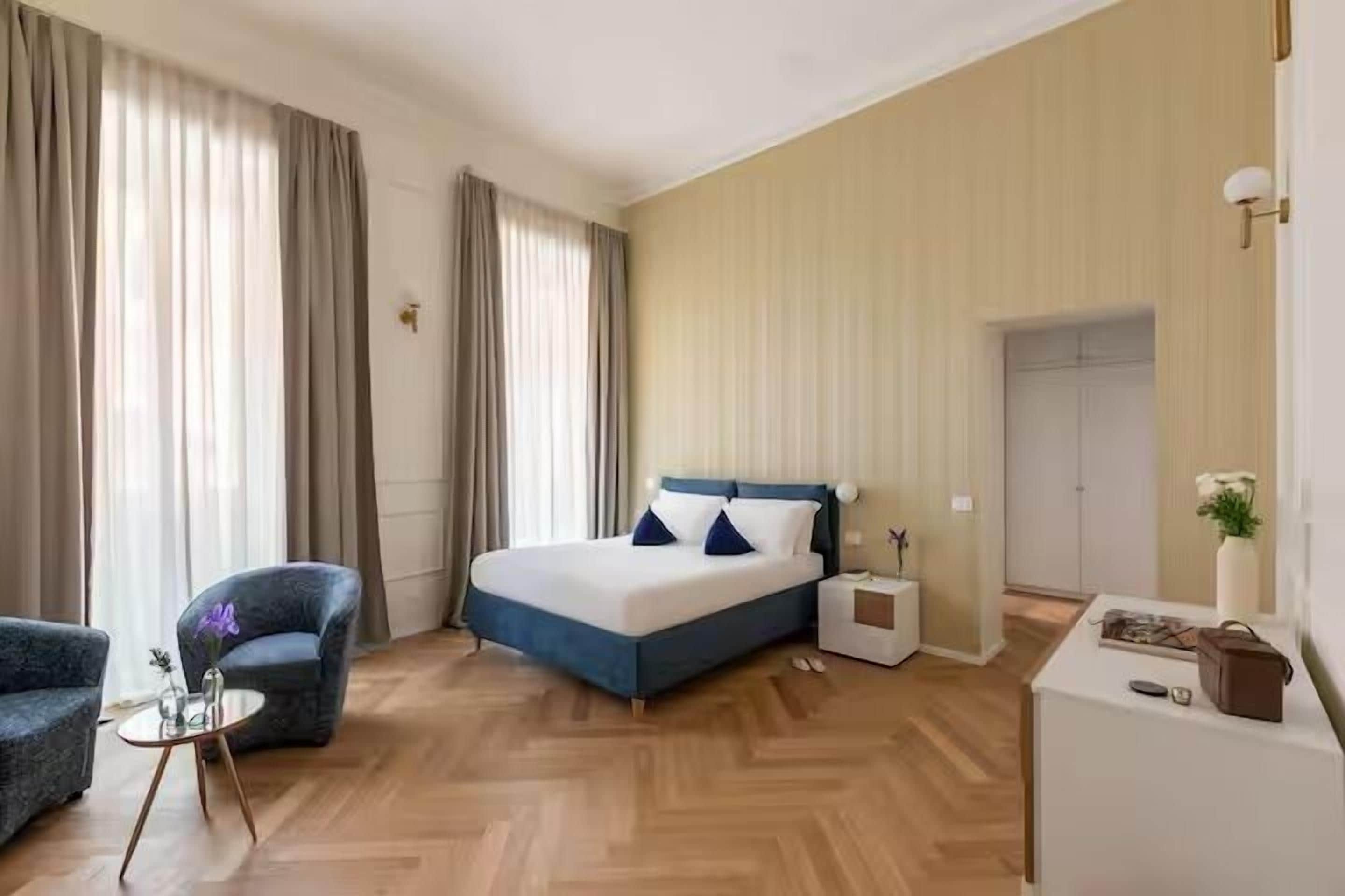 A hotel room with a bed, a chair, a small table, a nightstand, and a desk.