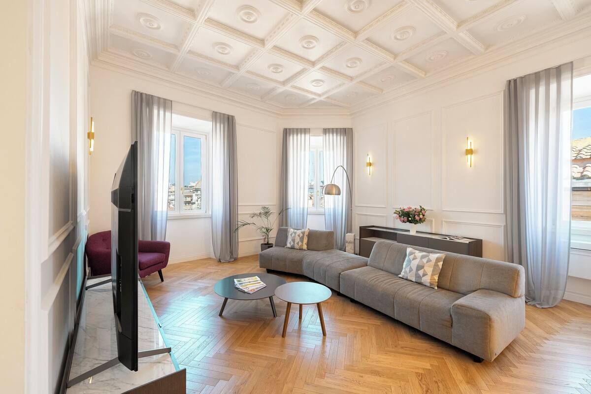Een ruime woonkamer met een grote grijze bank, twee ronde salontafels, een flatscreen televisie en een stadszicht door grote ramen.