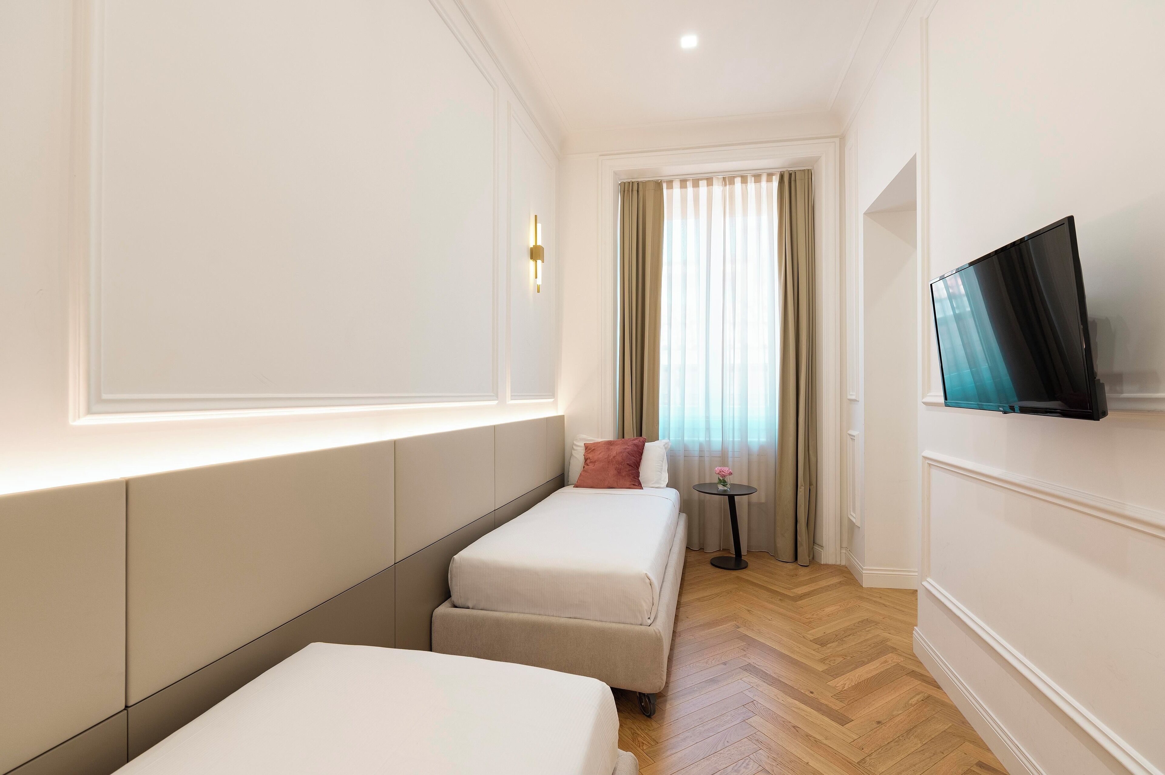 A hotel room with two single beds, a TV, a small table with a vase, and a window with curtains.