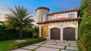 A two-story house with a circular tower, two garage doors, and a well-maintained garden with a palm tree.