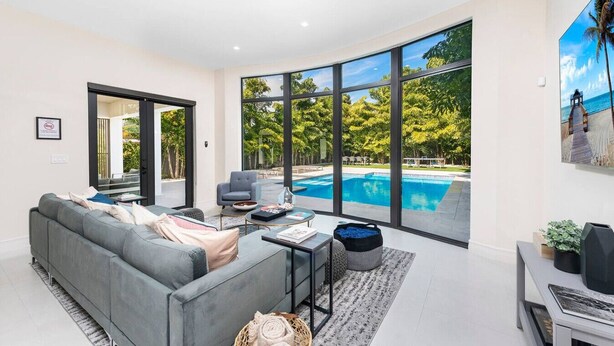 A modern living room with a grey sofa, a coffee table, and a large window overlooking a swimming pool and lush greenery.