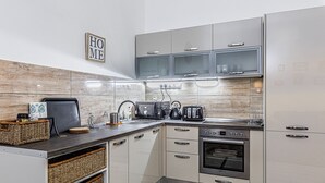 Une cuisine moderne avec des armoires blanches, un four encastré, un four à micro-ondes et un grille-pain. On trouve un panier en osier sur le comptoir et une enseigne « HOME » au mur.