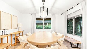 Un comedor moderno con una gran mesa de madera, sillas blancas y un banco. Hay dos mesitas de noche con lámparas y un amplio ventanal con cortinas.