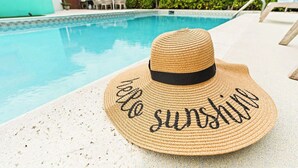 Un sombrero de paja con la frase 'hello sunshine' bordada, colocado junto a una piscina con agua azul cristalina y un suelo de baldosas.