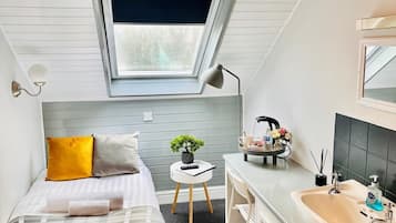 A compact bedroom with a skylight, a bed with a plaid cover, a small desk, and a bathroom with a sink and mirror.