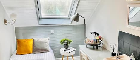 A compact bedroom with a skylight, a bed with a plaid cover, a small desk, and a bathroom with a sink and mirror.