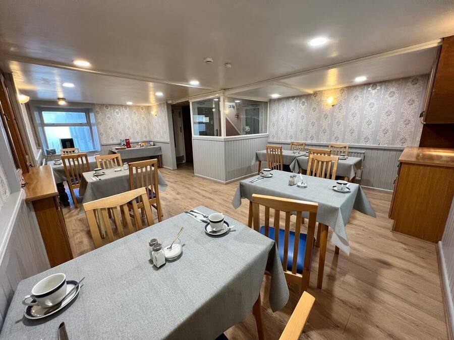 Un comedor con mesas dispuestas para dos, sillas de madera y vistas a la cocina a través de un separador de vidrio.
