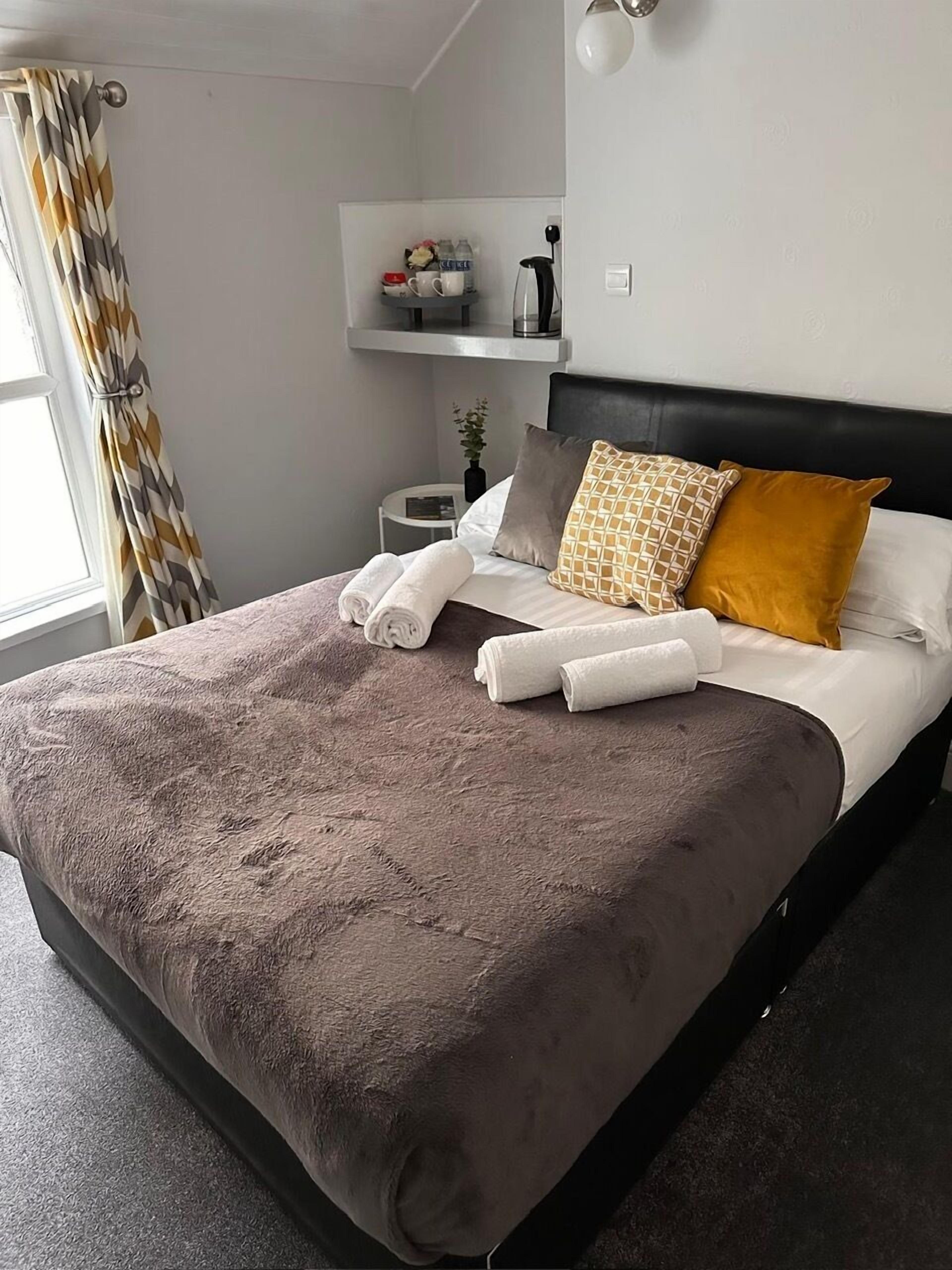 Un lit bien fait, avec une couverture marron et des oreillers, une table de chevet avec une lampe, et une étagère au-dessus du lit où sont rangés les articles de toilette.
