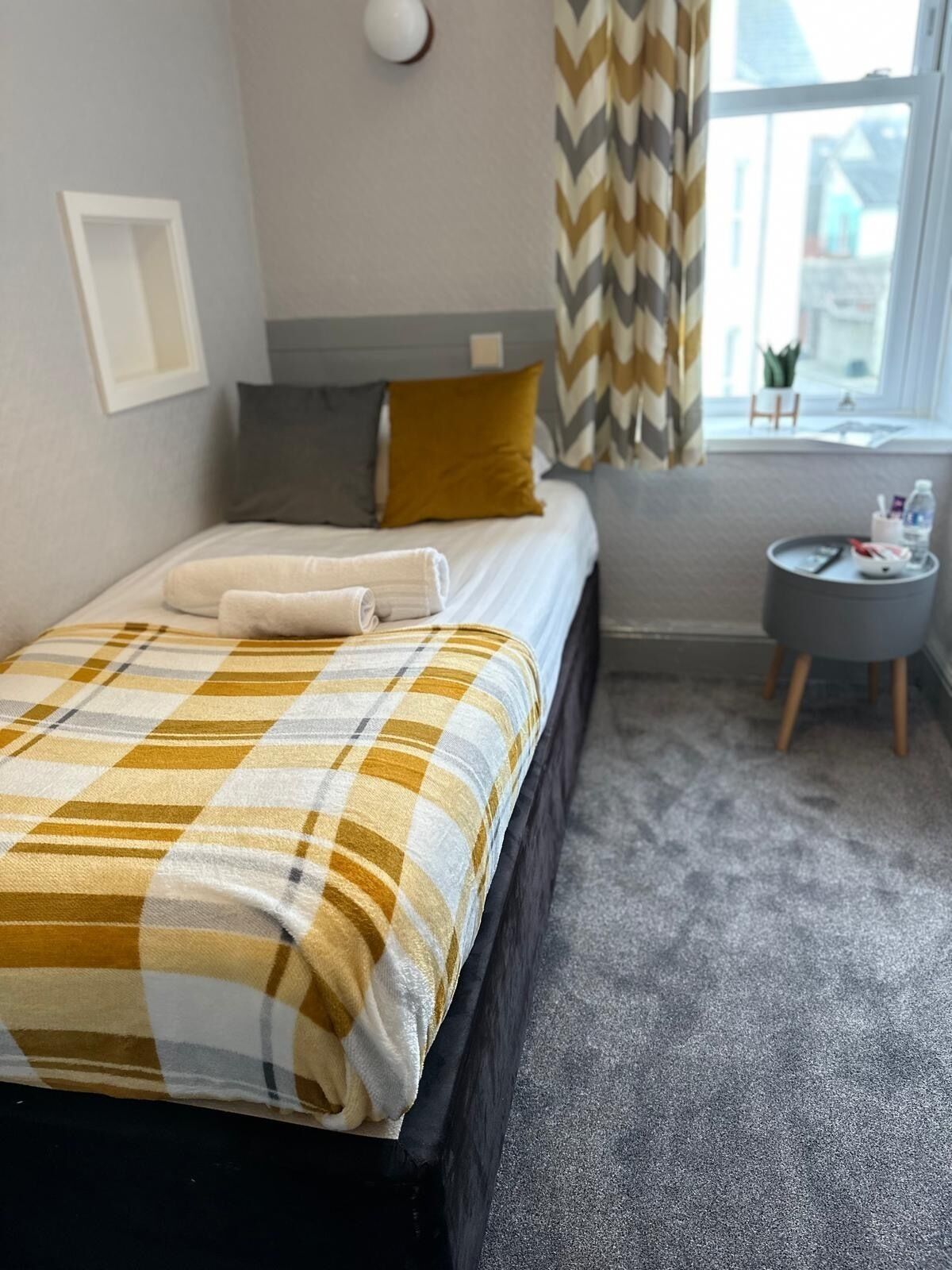 A neatly made bed with a plaid blanket, a grey headboard, and a small bedside table with a lamp and a bottle.