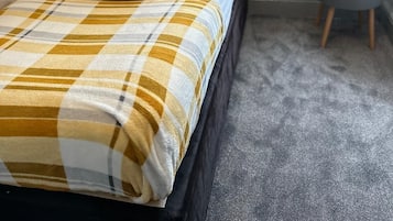 Una cama bien hecha con una manta escocesa, un cabecero gris y una mesita de noche pequeña con una lámpara y una botella.