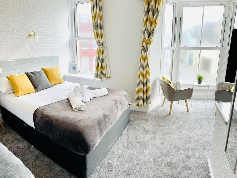 Une chambre d’hôtel moderne avec un grand lit, deux fauteuils, une petite table avec une plante et de grandes fenêtres équipées de rideaux.