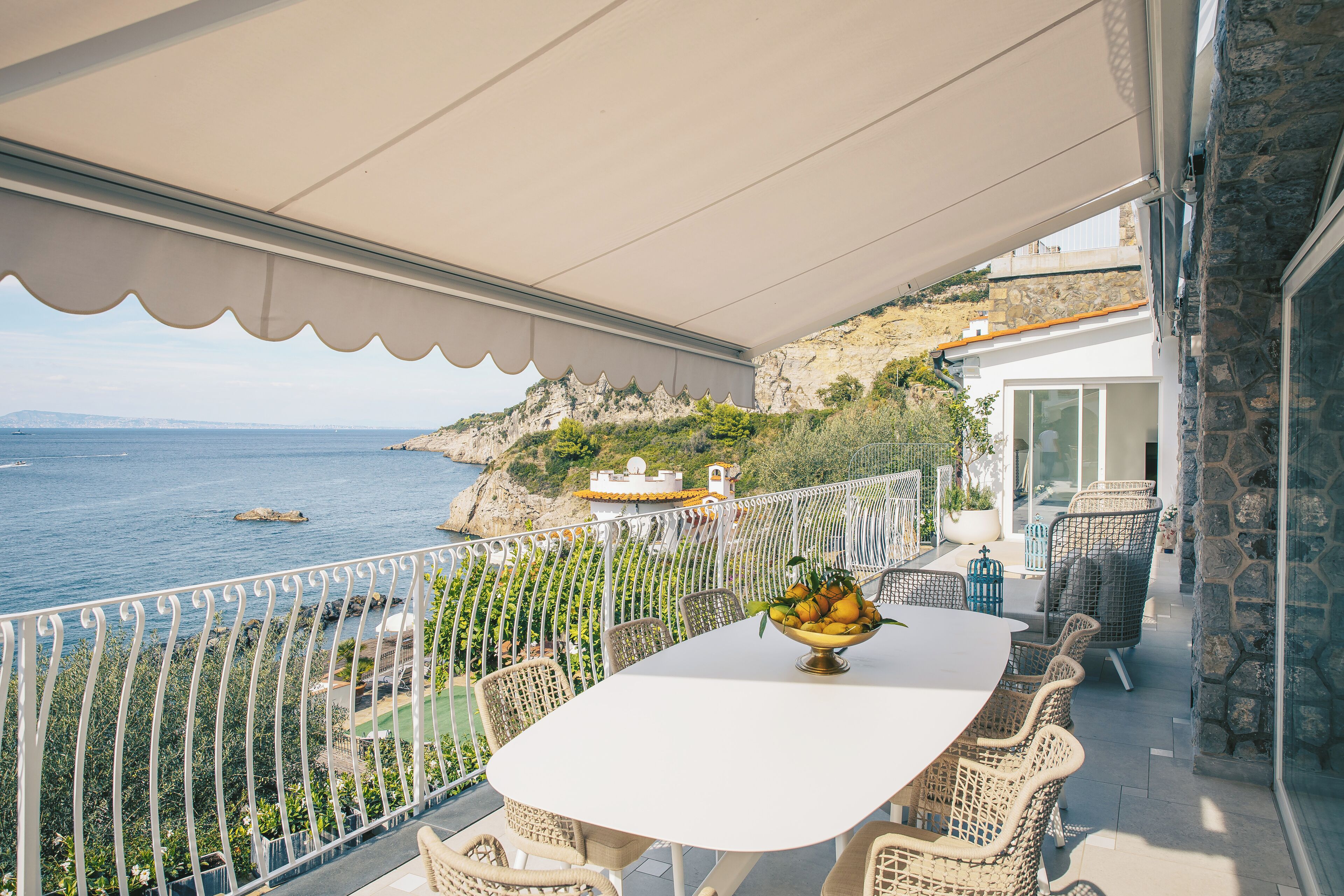 A balcony with a white table and wicker chairs, overlooking a coastal landscape with a view of the sea and cliffs.