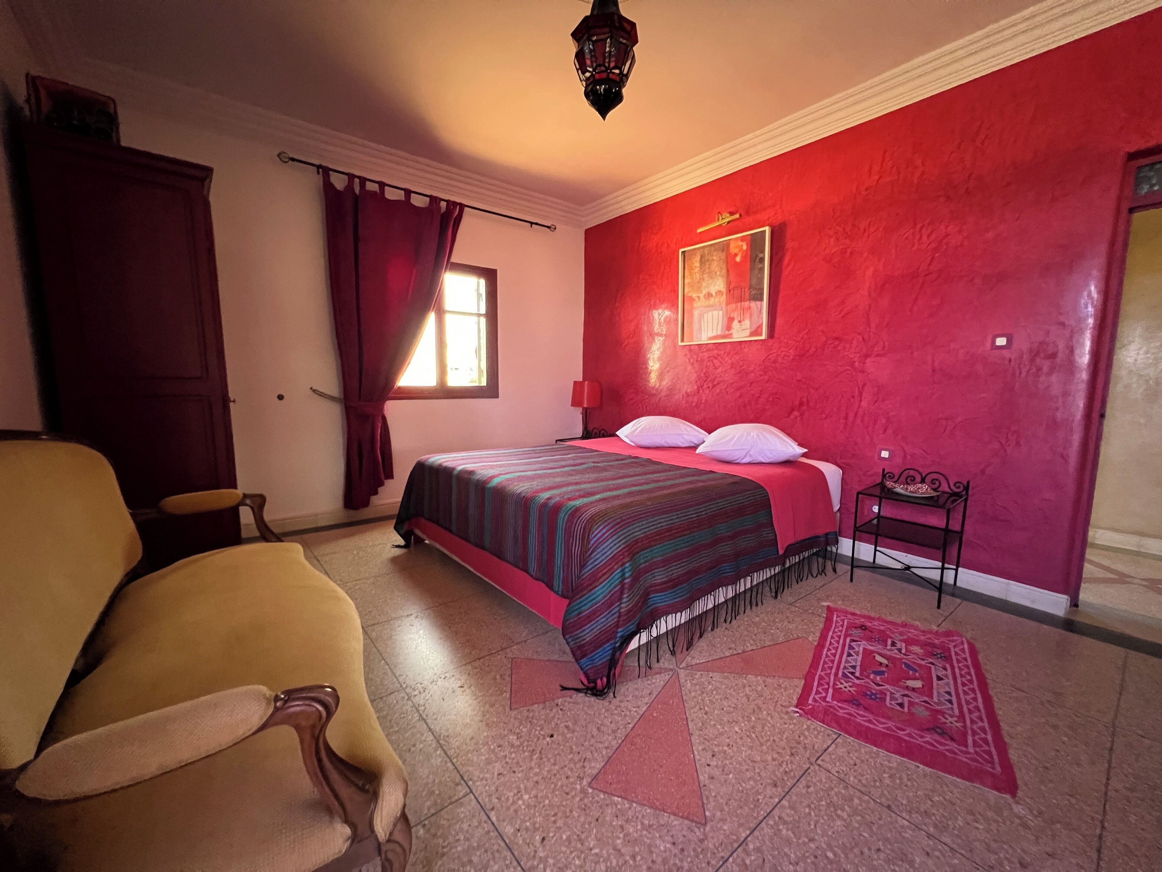Une pièce avec un lit, une chaise, une fenêtre avec des rideaux, un mur rouge et un tapis à motifs.