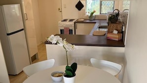 A modern kitchen with white cabinets, a refrigerator, and a stove. There is a dining area with a round table and chairs, and a potted plant on the table. - Beach Getaway Villa: 2BR Fun! (Bicton)