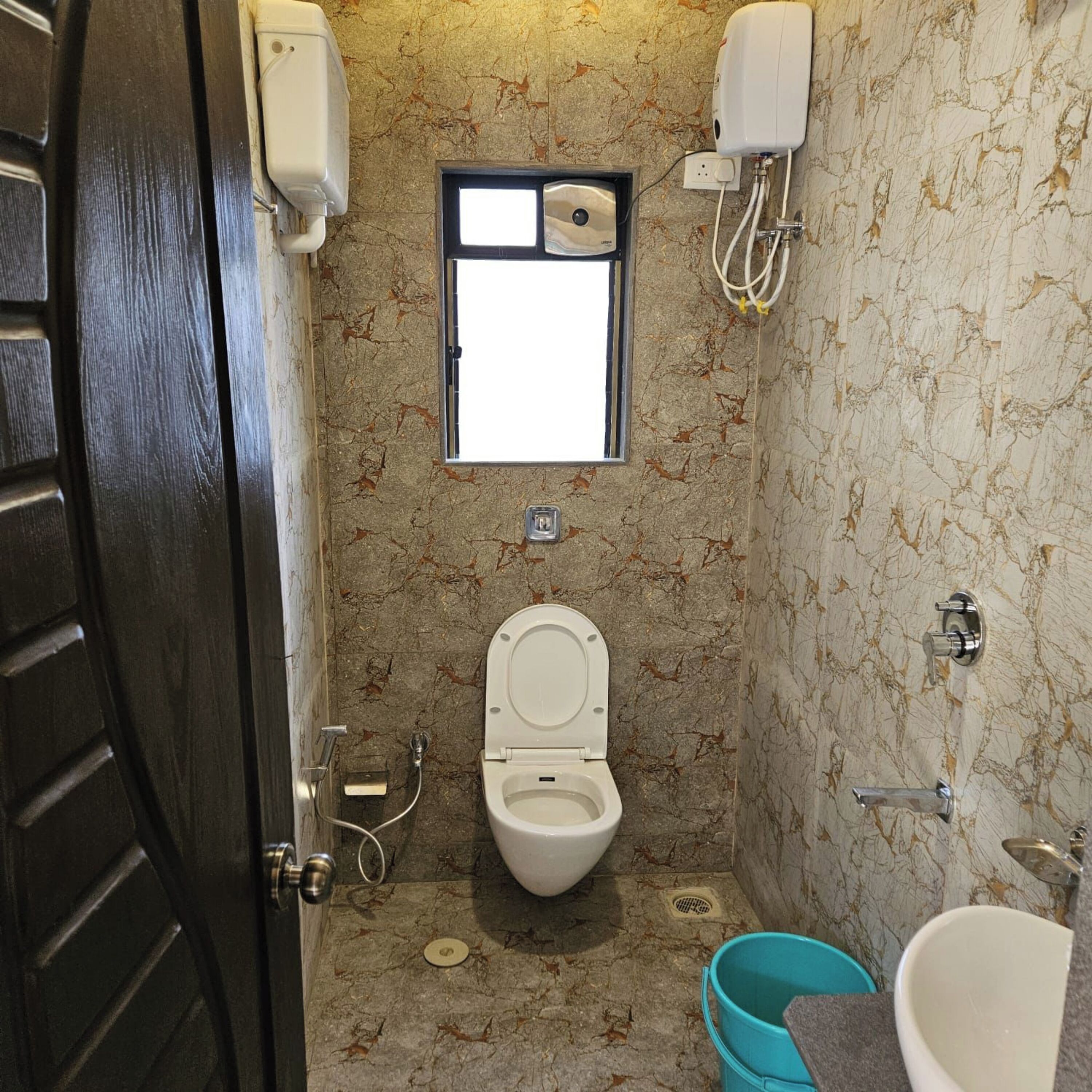 A small bathroom with a toilet, a window, a hand dryer, and a bucket.
