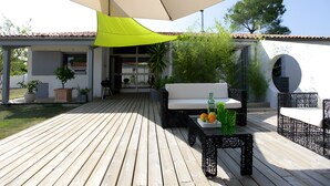 A wooden deck with outdoor furniture, a white sofa, and a table with fruits and bottles. - Spacious Villa 10 min from Royan – 4 Bedrooms, Garden, Terrace, Ideal for Families and Friends (Saujon)