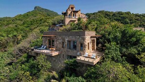 A stone villa with a balcony and outdoor seating area, surrounded by lush greenery and a mountain backdrop.