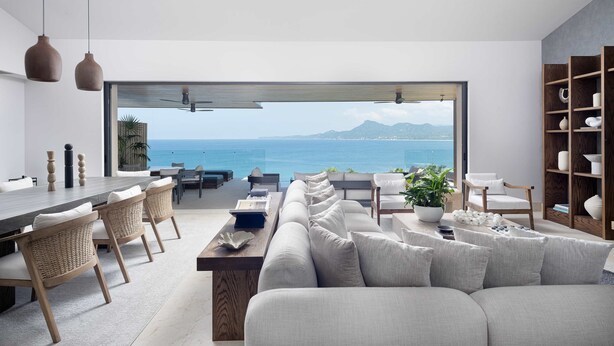A modern living room with a large window overlooking the sea, a dining area with wicker chairs, and a wooden bookshelf. - Casa Ravello: Luxury Villa, Full Service & Premier Golf (Punta Mita)
