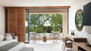 Un dormitorio moderno con una cama grande, un escritorio y dos sillas. Hay un balcón con vistas a árboles y barandilla de madera.