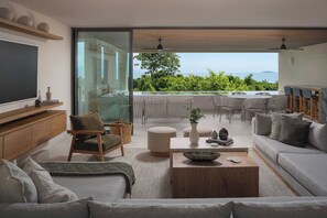 Ein modernes Wohnzimmer mit Sofa, Sessel und Couchtisch, einem Fernseher und Meerblick durch große Fenster.