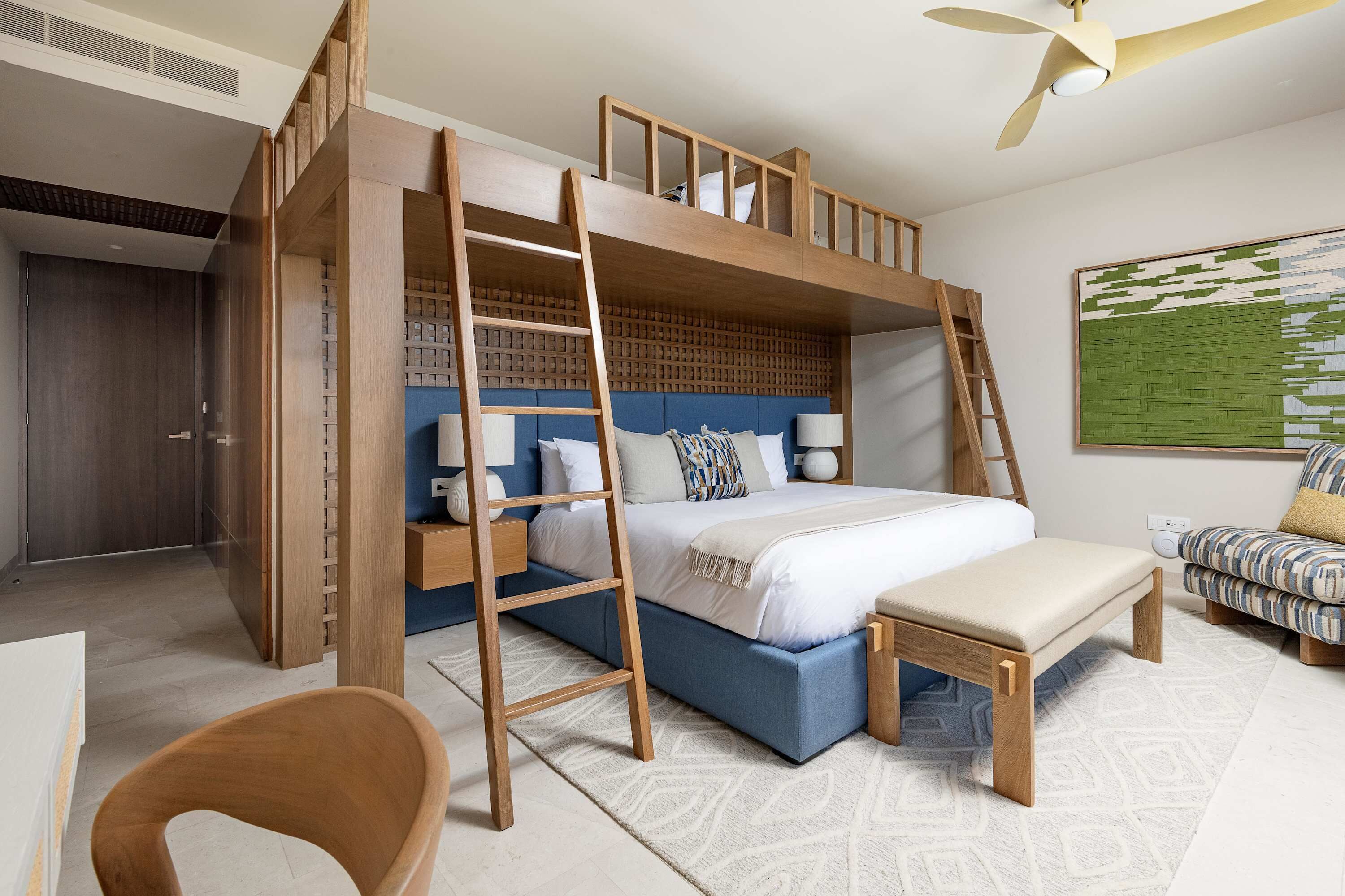 Un lit superposé avec une échelle, un bureau, une chaise et un canapé dans une pièce dotée d’un tapis à motifs et d’un ventilateur de plafond.
