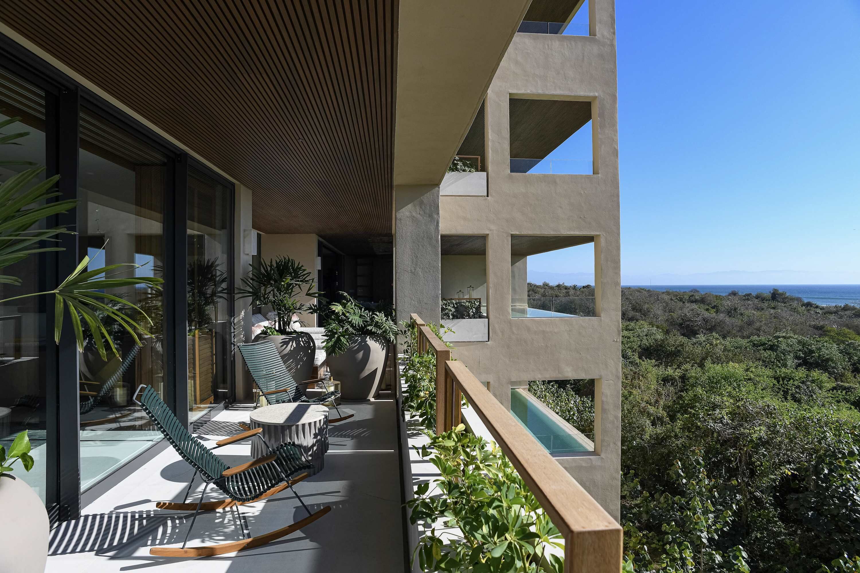 Een modern balkon met uitzicht op de oceaan, voorzien van stoelen en een tafel, omgeven door groen.