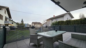 An outdoor patio with wicker furniture and a glass table, surrounded by a metal fence and houses in the background. - FeWo in der Kiese Hard - Top 2 "Bodensee" with a large terrace (Hard)