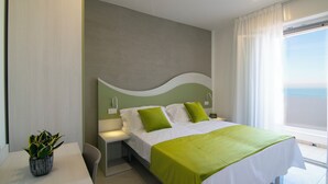 A modern hotel room with a large bed, green pillows, a bedside table with a plant, and a view of the sea through the window. - Residence Flow-r (Riccione)