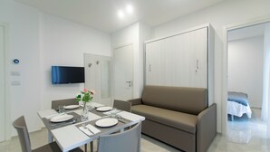 A modern, well-lit room with a dining table set for two, a sofa, a TV mounted on the wall, and a wardrobe. - Residence Flow-r (Riccione)
