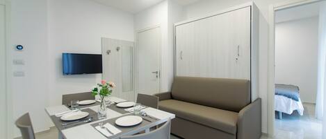 A modern, well-lit room with a dining table set for two, a sofa, a TV mounted on the wall, and a wardrobe.
