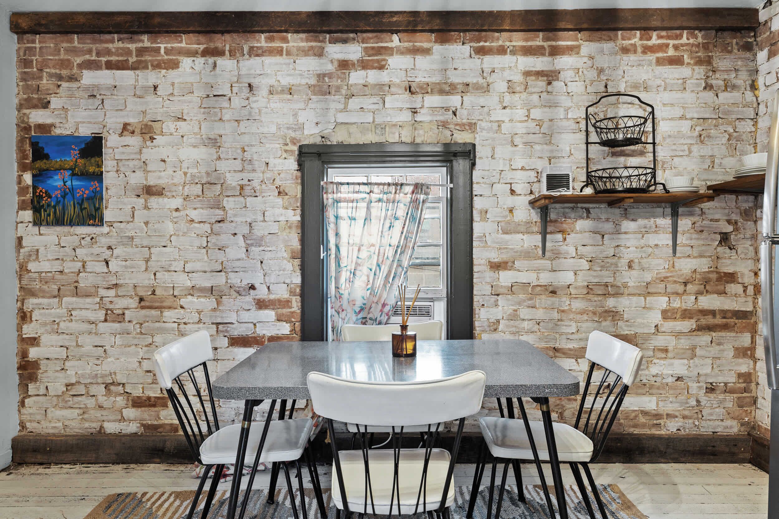 A dining area with a brick wall, a window with curtains, a metal basket shelf, and a painting.