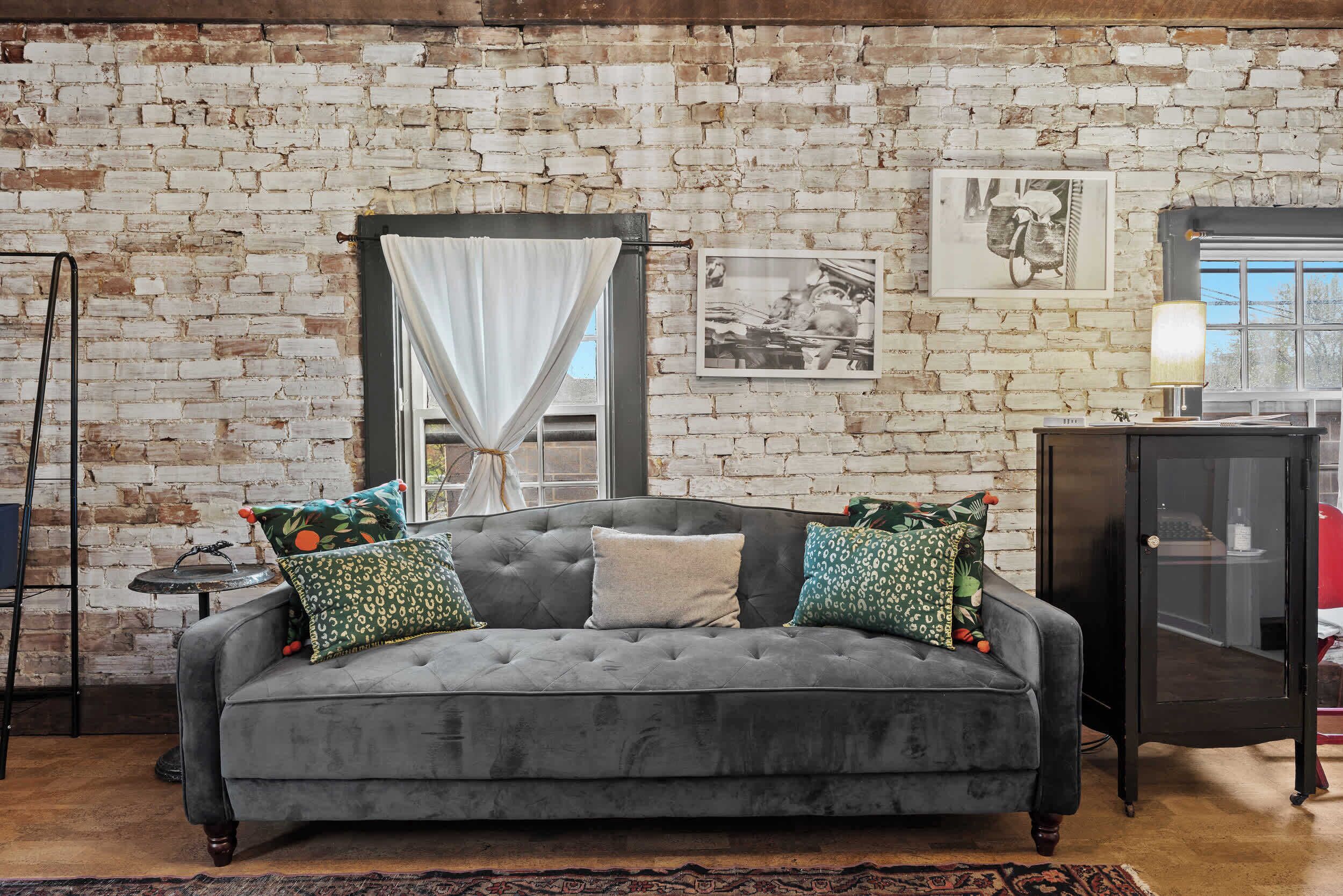 A grey sofa with patterned cushions, a brick wall, a window with white curtains, and a framed picture on the wall.