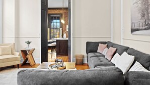 A living room with a grey sofa, a wooden side table, and a beige armchair. There is a kitchen visible through an open door in the background. - Classic & elegant Victorian home in Old Louisville (Louisville)