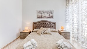 A neatly made bed with a headboard, two bedside tables with lamps, a decorative wall piece, and a window with sheer curtains. - Apartment 'Mirea' with Private Terrace, Wi-Fi and Air Conditioning (Melissano)