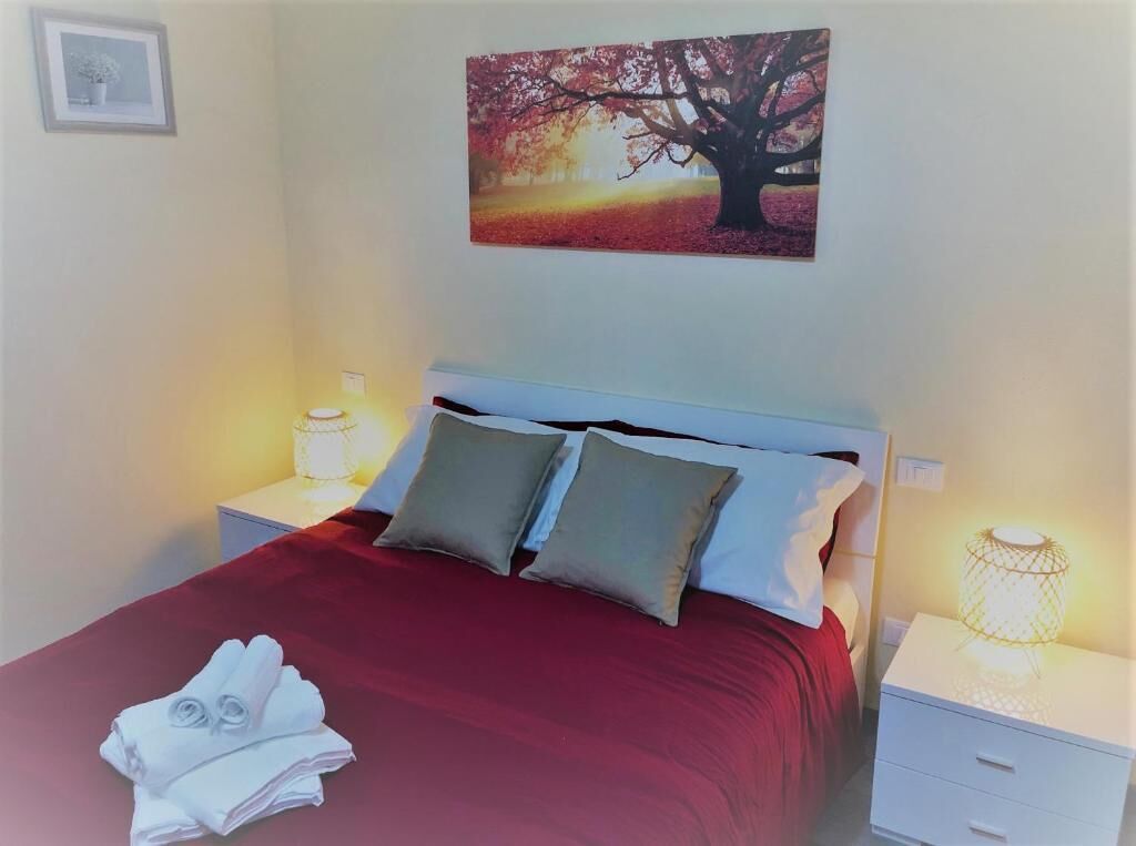 Una cama bien hecha con una colcha roja y dos almohadas, una lámpara de noche y un cuadro con un árbol en la pared.