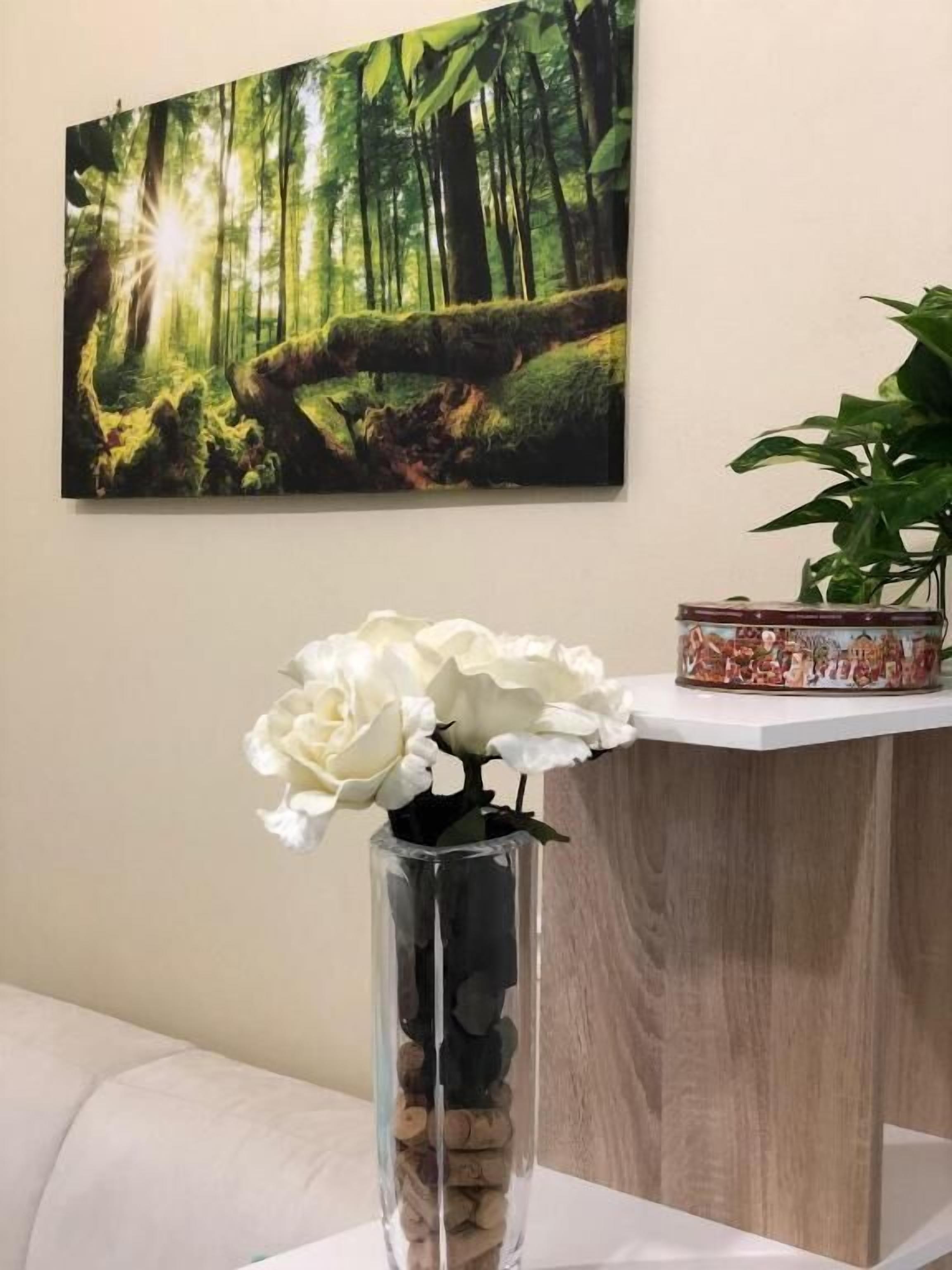 A room with a painting of a forest, a vase with white flowers, and a plant on a shelf.