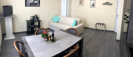 Una sala de estar moderna con una mesa de comedor, sofá y sillas. Hay un televisor sobre un mueble bajo, una cafetera y una mesita con una planta.