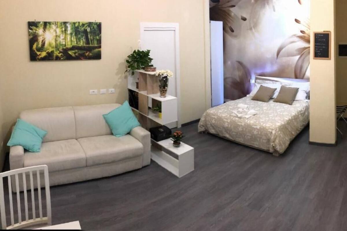 A compact hotel room with a sofa, a bed, a small side table, and a wall-mounted shelf with decorative items.