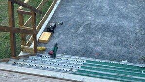 Et træterrasse med gelænder af metal, et grønt elværktøj og byggematerialer på en grusoverflade.