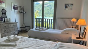 Et hotelværelse med to senge, en kommode, en stol, et bord og en balkon med udsigt over grønne bakker.