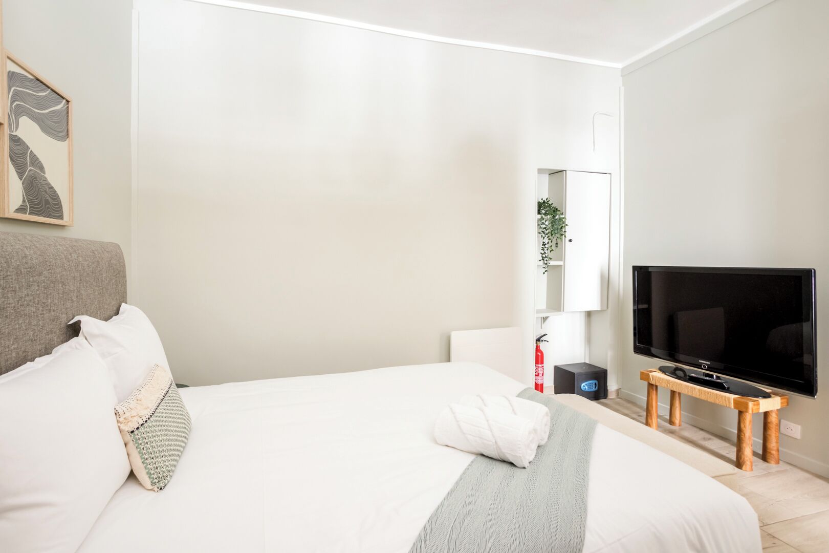 Et moderne hotelværelse med en stor seng, et fladskærms-tv på et træstel, et hvidt skab og en lille plante.
