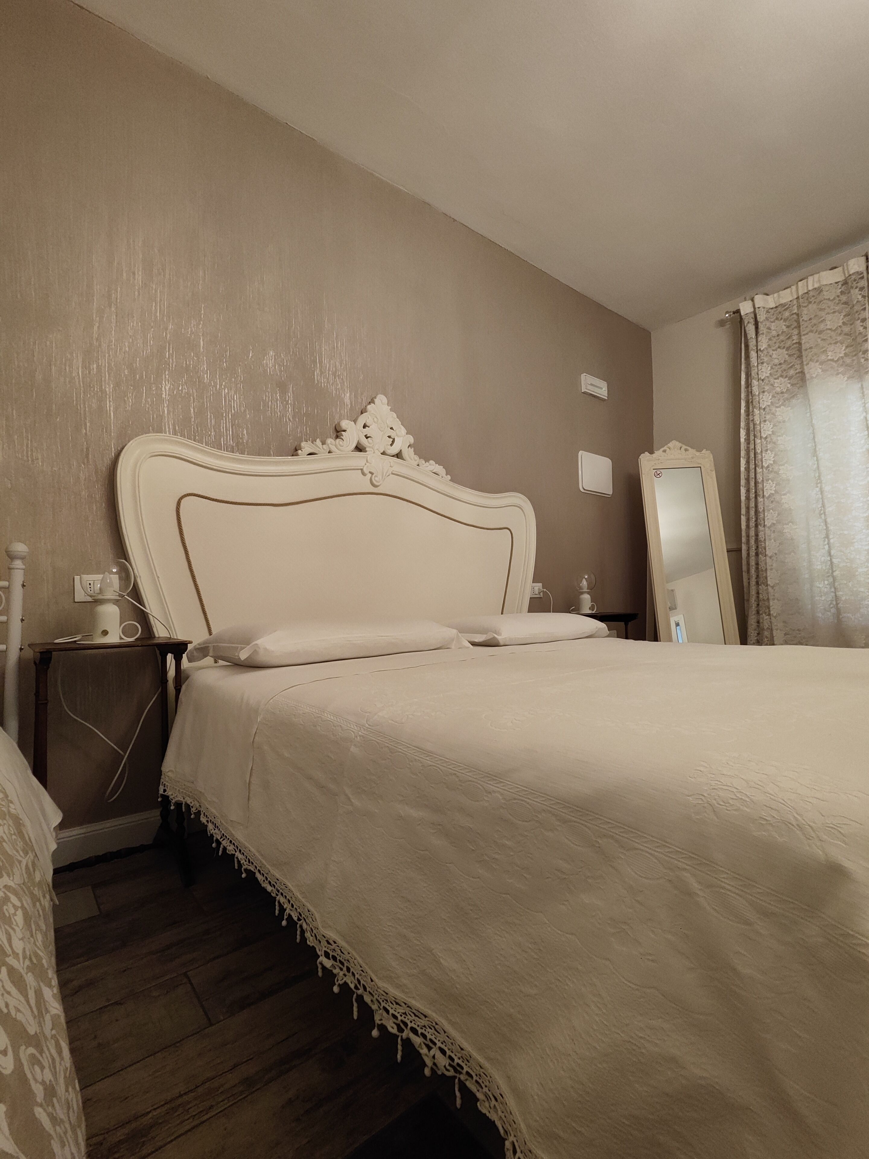 A bedroom with a large bed, a nightstand, a bedside table, a mirror, and a window with curtains.