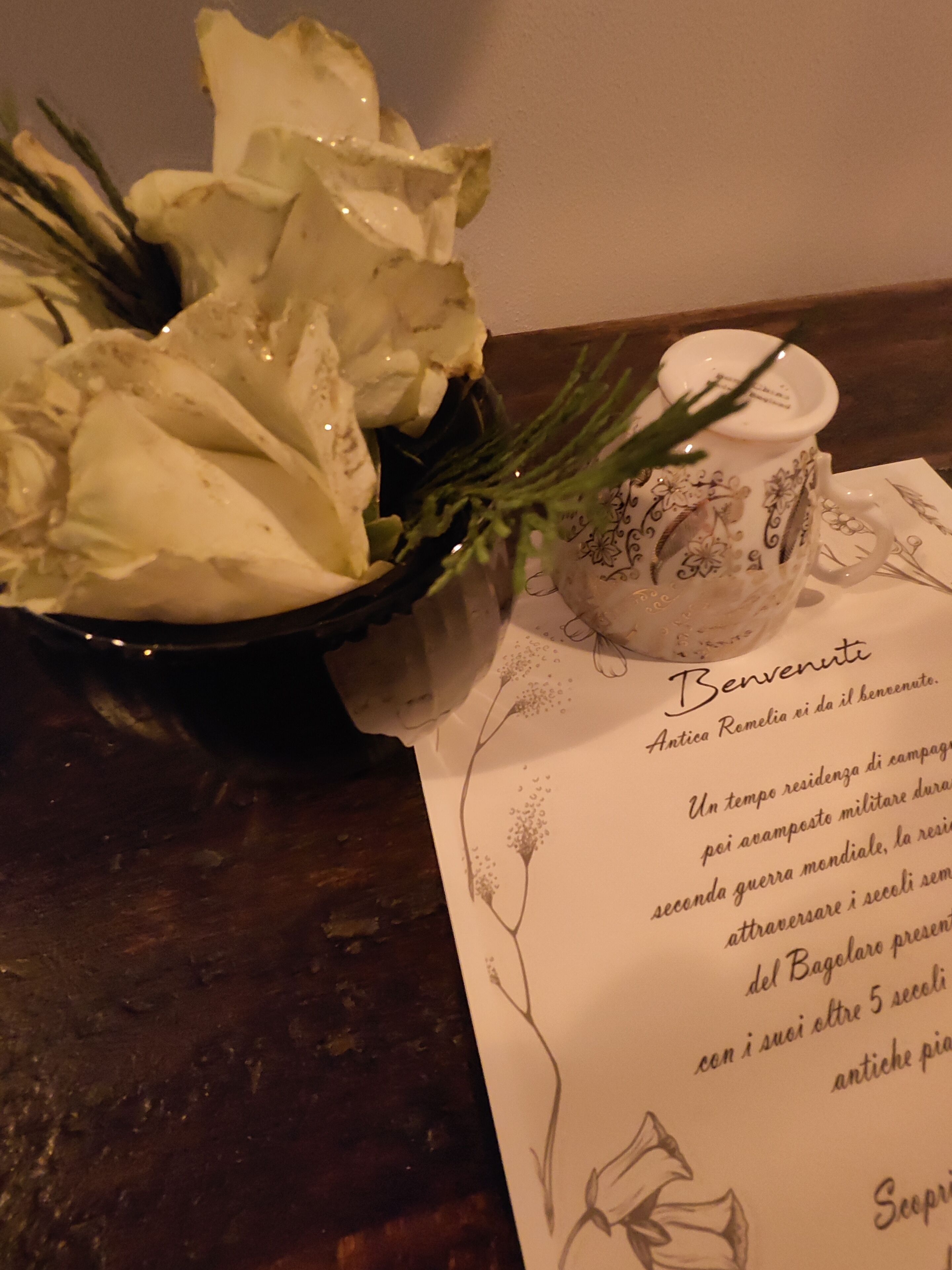 A table with a bowl of white flowers, a ceramic mug with floral designs, and a menu with Italian text.