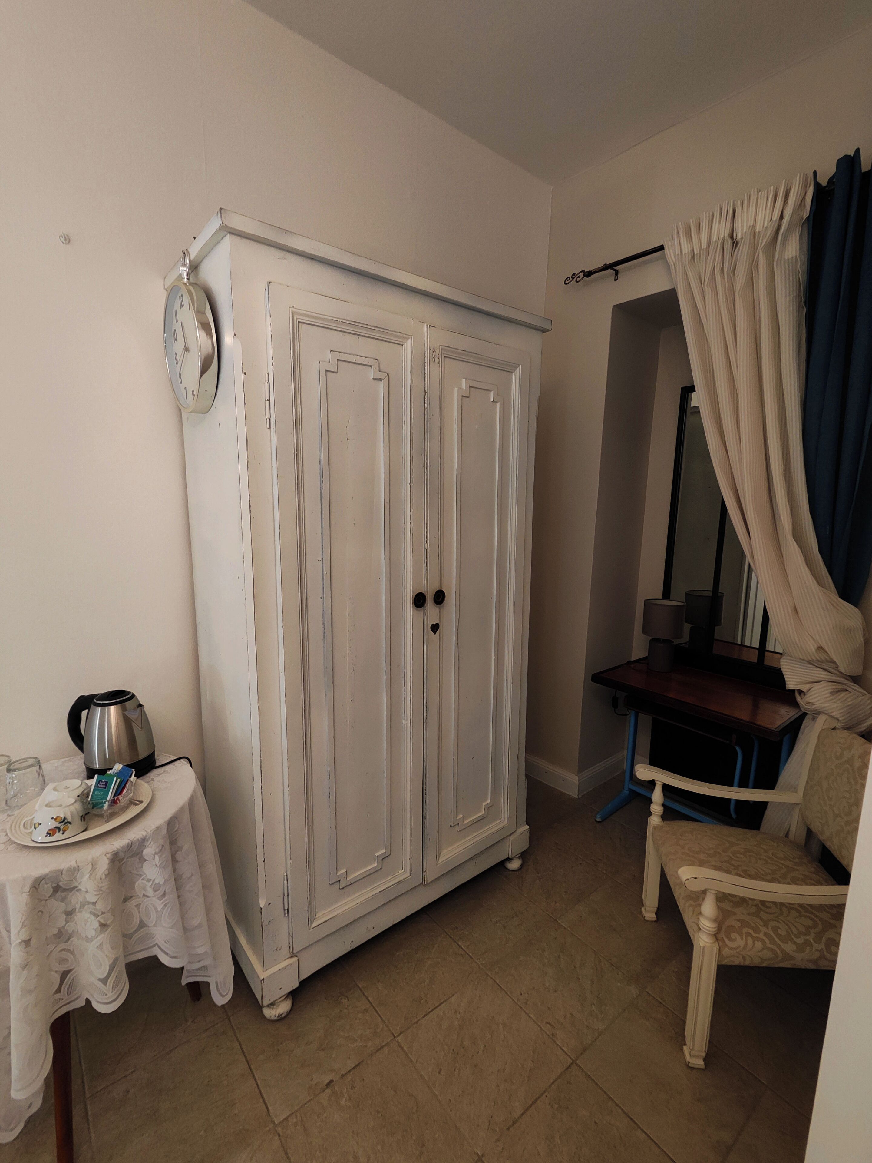 Een kamer met een witte kast, een klein tafeltje met waterkoker en kopjes, een stoel en een spiegel met gordijnen.