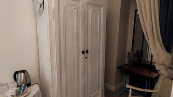 A room with a white wardrobe, a small table with a kettle and cups, a chair, and a mirror with curtains.